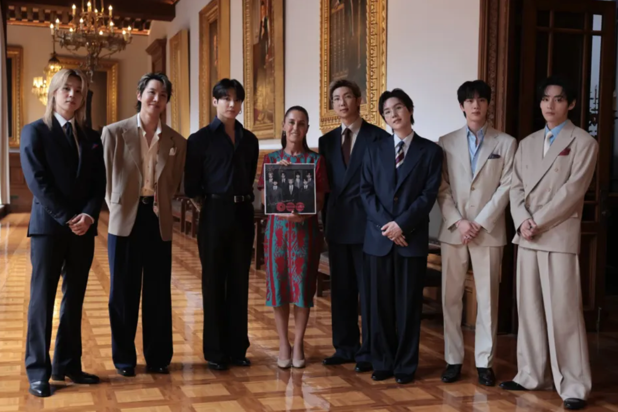 Los siete integrantes de BTS — RM, Jin, Suga, J-Hope, Jimin, V y Jung Kook — saludando desde el balcón central del Palacio Nacional de México junto a la presidenta Claudia Sheinbaum, mientras decenas de miles de fanáticos con luces moradas llenan el Zócalo de la Ciudad de México al fondo en una tarde soleada.